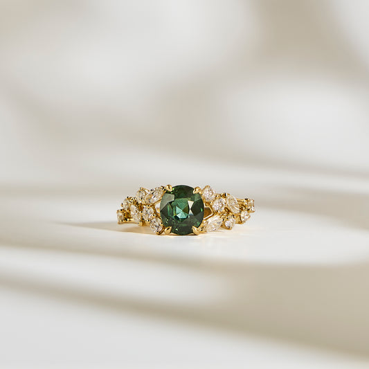 La Bague Saphir Teal • 2,07ct de Bonnot Paris, une bague en or avec un centre en saphir bleu sarcelle saisissant entouré de pierres claires, repose élégamment sur une surface crème doucement éclairée avec des ombres douces.
