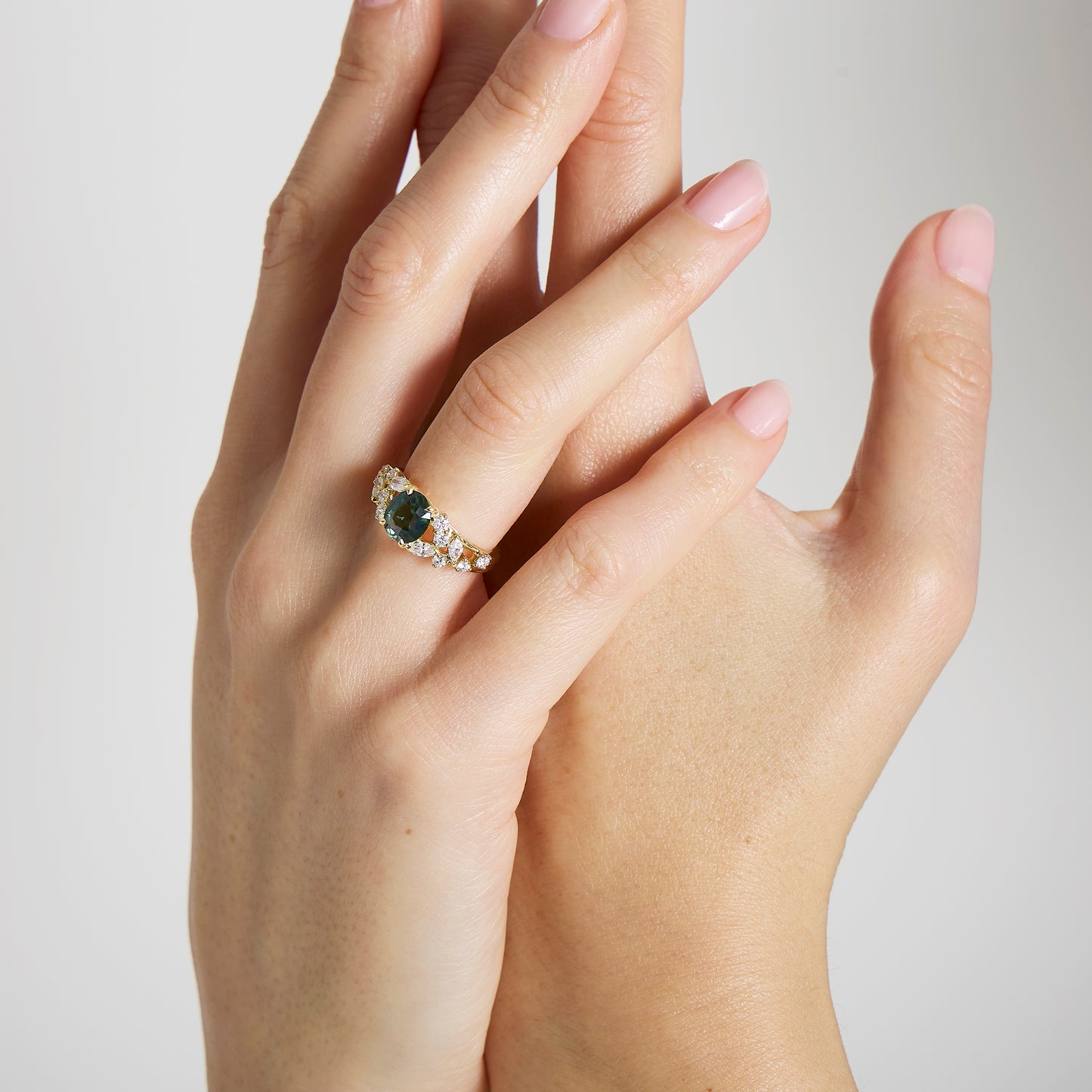 Gros plan sur deux mains claires ; l'une est ornée d'une bague Bonnot Paris Bague Saphir Teal • 2,07 ct ornée d'un saphir turquoise encadré de diamants taille marquise. Les ongles sont manucurés avec un vernis rose clair sur un fond sobre.
