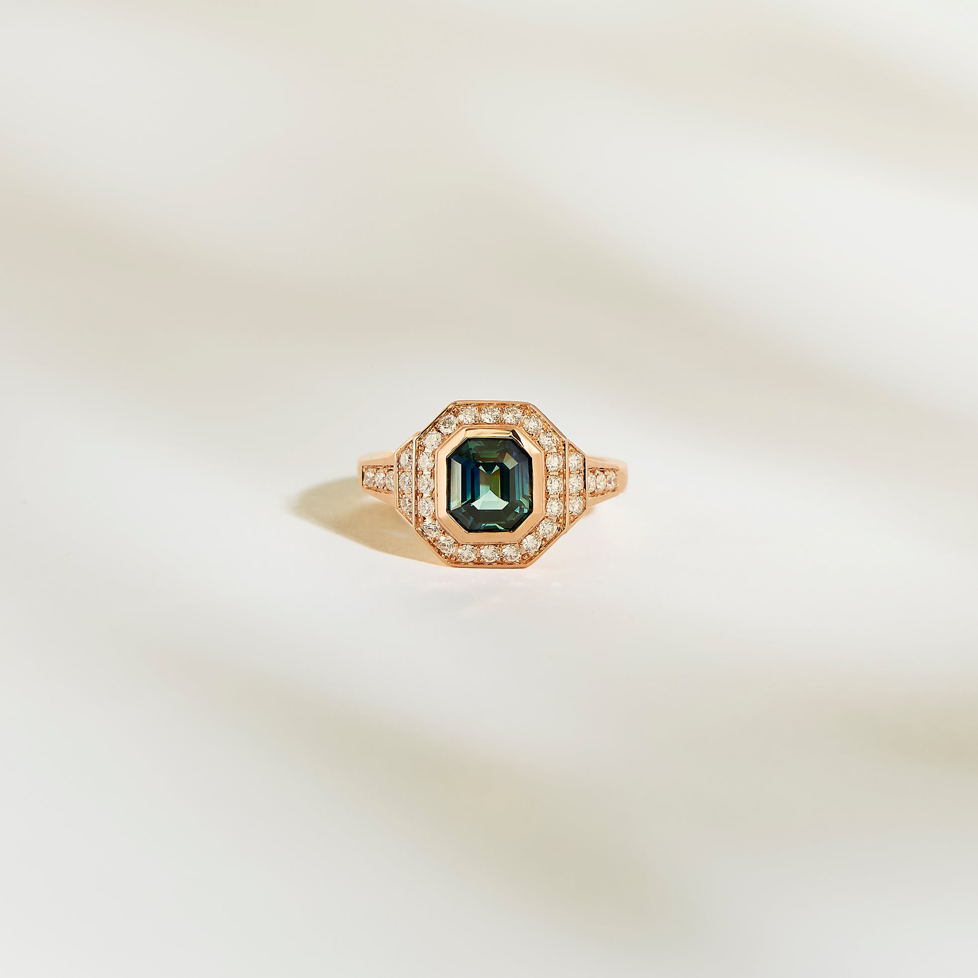La Bague Saphir Teal de Bonnot Paris, une bague en or ornée d'une pierre précieuse vert foncé taille émeraude de 1,56 ct entourée d'un halo de petits diamants blancs, est élégamment mise en valeur sur un fond doux et clair.
