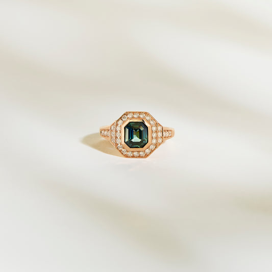 La Bague Saphir Teal de Bonnot Paris, une bague en or ornée d'une pierre précieuse vert foncé taille émeraude de 1,56 ct entourée d'un halo de petits diamants blancs, est élégamment mise en valeur sur un fond doux et clair.