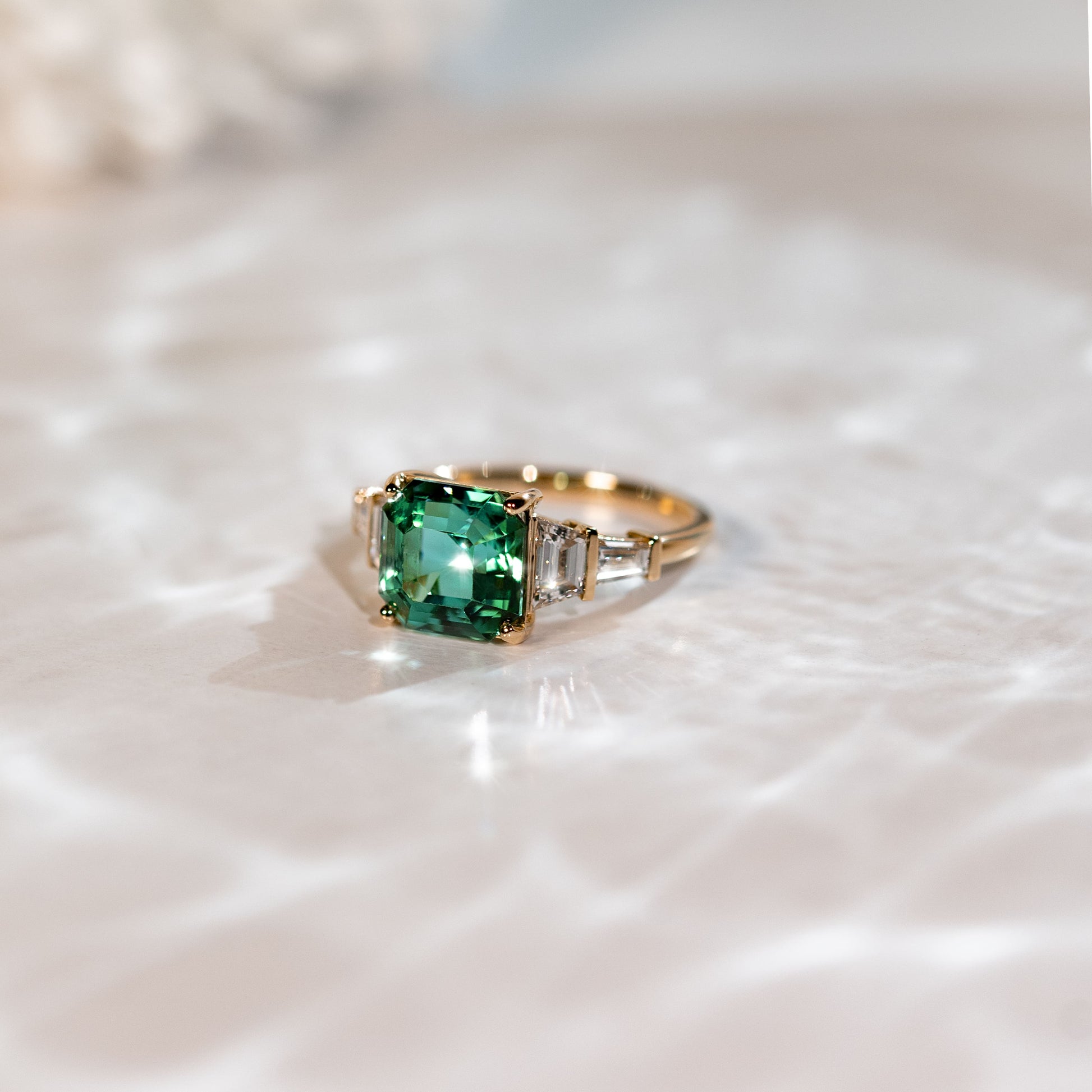 La Bague Tourmaline · 4,35ct de [NOUVEAU] Bonnot Paris présente une bague en or avec une grande pierre centrale carrée en émeraude et deux diamants taille baguette plus petits, présentés sur une surface réfléchissante blanche qui projette des ombres et de la lumière subtiles.