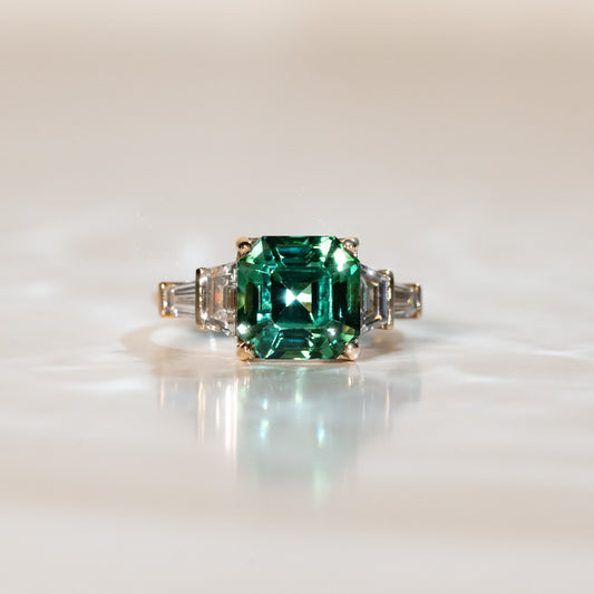 La Bague Tourmaline · 4,35 carats de [NOUVEAU] Bonnot Paris met en valeur une tourmaline verte taille carrée au centre, flanquée de diamants baguette plus petits. Présentée sur une surface lisse et réfléchissante, elle respire l'élégance intemporelle de l'Art Déco.