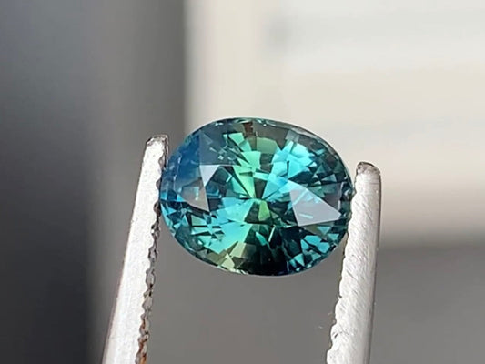 Une pierre précieuse ronde et facettée de la collection « Saphir Teal • 1,6 ct » de Bonnot Paris est maintenue par une pince métallique. Sa surface scintille de nuances de bleu et de touches de vert sur un fond neutre et flou.