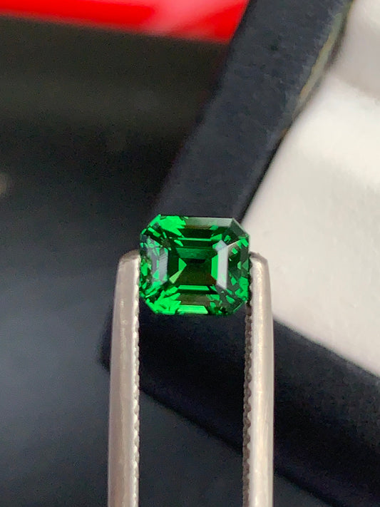 Une paire de pinces tient la Tsavorite Bonnot Paris • 1,57 ct, sa teinte verte vibrante brillant sur le fond sombre, avec une partie d'un support de bague subtilement visible.
