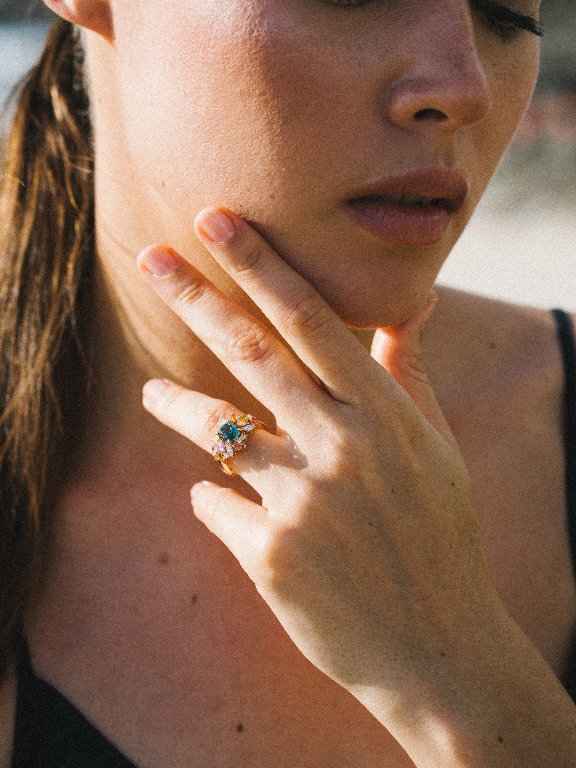 Une personne à la peau claire pose son menton sur sa main, mettant en valeur la bague Sea Bloom - Tourmaline de Bonnot Paris. Ses longs cheveux bruns sont attachés en arrière et elle porte un haut noir. L'image est doucement éclairée par la lumière naturelle, créant une atmosphère sereine.