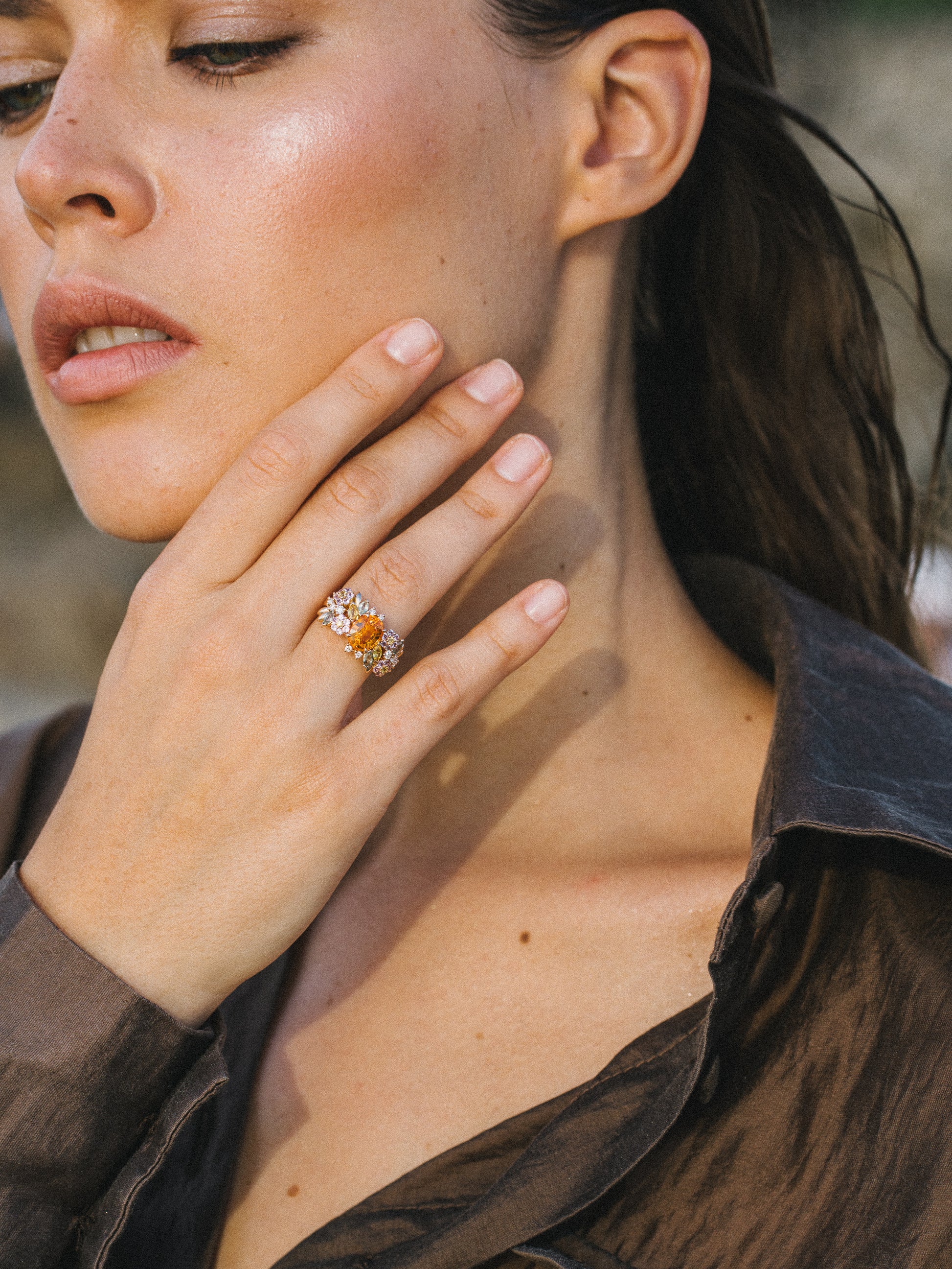 Une personne, cheveux mouillés et maquillage léger, se touche le visage, mettant en valeur une bague ornée de pierres précieuses qui rappelle l'élégance du Golden Bloom – Grenat Mandarin de Bonnot Paris. Elle porte un chemisier sombre transparent sur un fond légèrement flou, suggérant un décor extérieur subtilement sophistiqué.