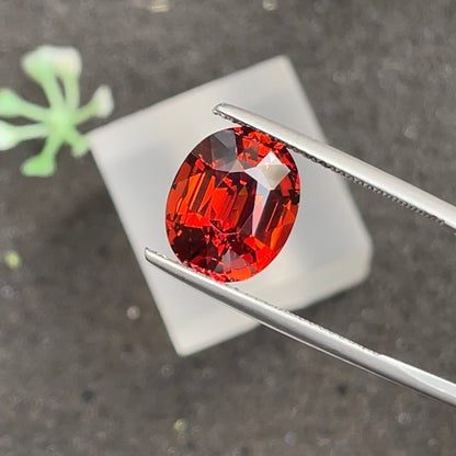 Une pierre précieuse vibrante Grenat Spessartite · 7,76 ct de Bonnot Paris est délicatement maintenue entre des pinces métalliques, sur un fond sombre avec des notes d'une plante verte floue et d'un objet carré blanc à proximité.