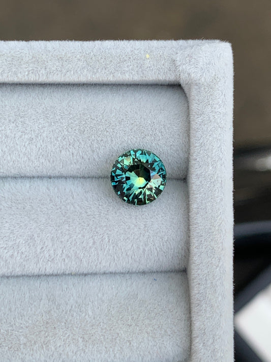 La pierre précieuse Bonnot Paris Saphir Teal • 2,55 carats de Madagascar scintille de reflets bleu sarcelle et aqua sur un fond gris doux, sa surface facettée évoquant l'élégance d'une exquise bague de fiançailles.