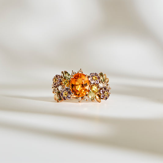 La bague Bonnot Paris Golden Bloom - Grenat Mandarin est ornée d'une pierre centrale ronde orange, probablement un grenat spessartite, entourée de pierres violettes et vertes éclatantes formant un motif floral. Sur un fond doux et lumineux, elle capture l'essence même de la Fleur de Couleur.