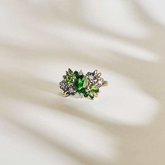 La bague Bonnot Paris Green Bloom - Tsavorite met en valeur une imposante pierre verte, entourée de saphirs bleus plus petits, de pierres incolores et de grenat tsavorite. Elle est élégamment présentée sur une surface crème doucement éclairée, projetant de subtiles ombres en arrière-plan.