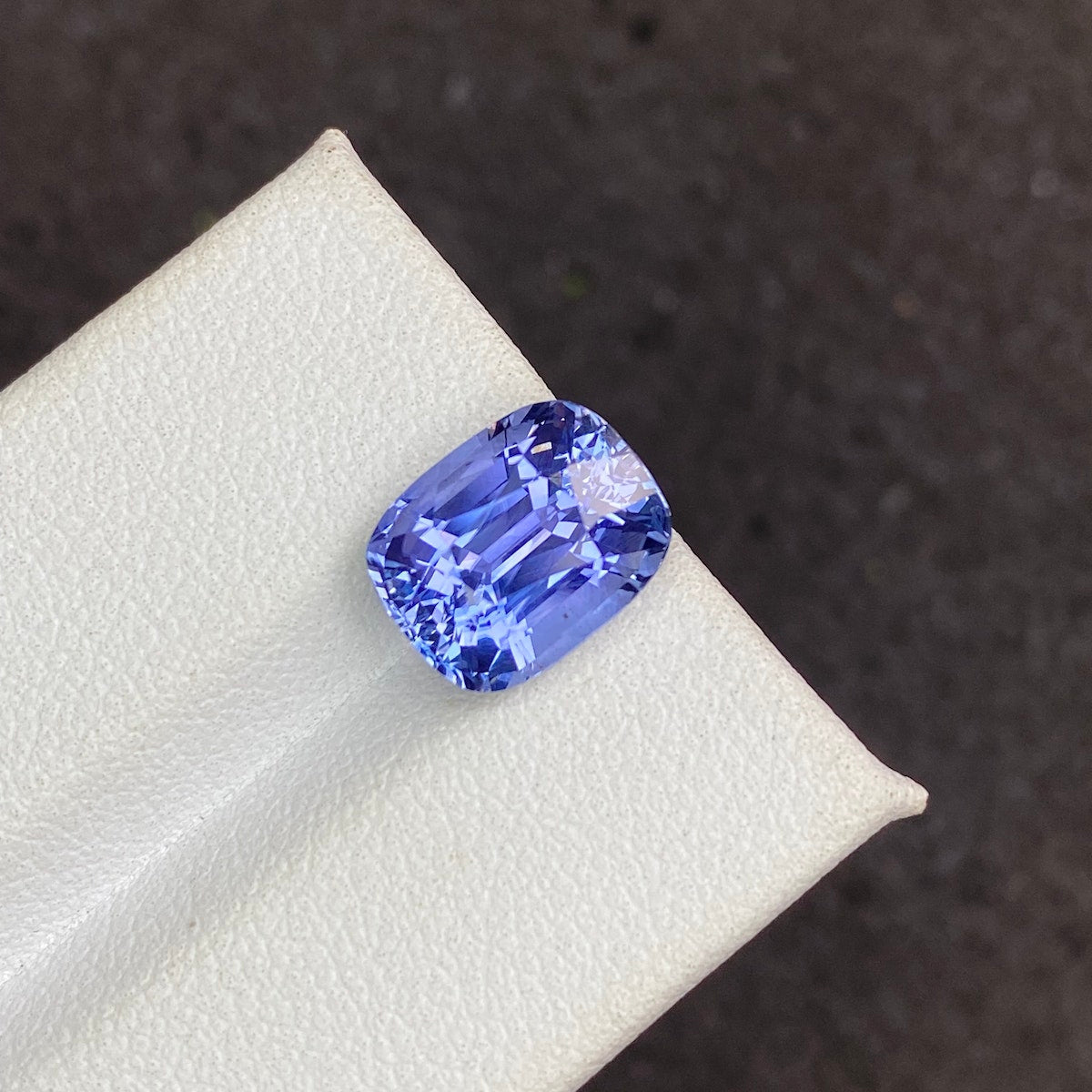 Une photo en gros plan présente le Saphir Bleu · 5,19 ct de Bonnot Paris, une pierre précieuse bleu vif taille coussin avec une surface facettée qui reflète brillamment la lumière, reposant sur une surface blanche texturée sur un fond sombre.