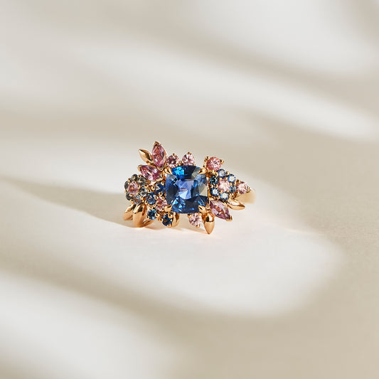L'exquis Navy Bloom - Saphir de Bonnot Paris présente un grand saphir bleu, orné de plus petites pierres roses et bleues dans un motif floral, reposant sur une surface crème qui rehausse sa beauté.