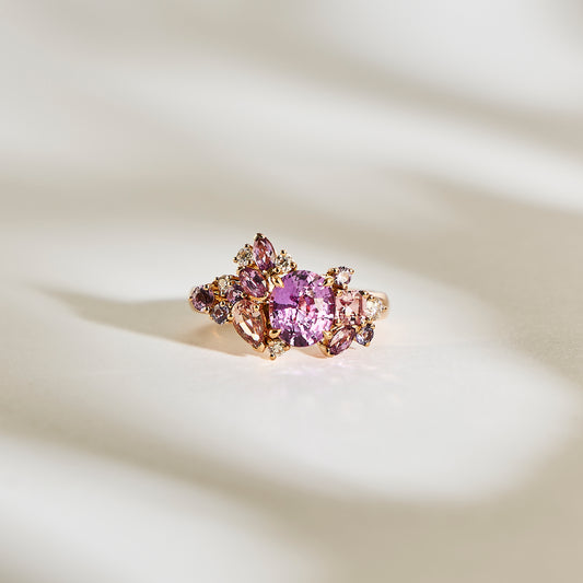 Le Purple Bloom - Saphir de Bonnot Paris présente une grande pierre précieuse rose entourée de diamants plus petits et de pierres claires, le tout serti sur une bande d'or, élégamment présentée sur une surface texturée doucement éclairée projetant des ombres douces.