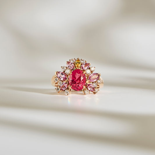 Le Rose Bloom - Spinelle Mahenge de Bonnot Paris présente une grande pierre précieuse rose ovale entourée de pierres plus petites taille marquise roses et claires dans un motif floral, le tout sur un fond doucement éclairé avec des ombres douces.
