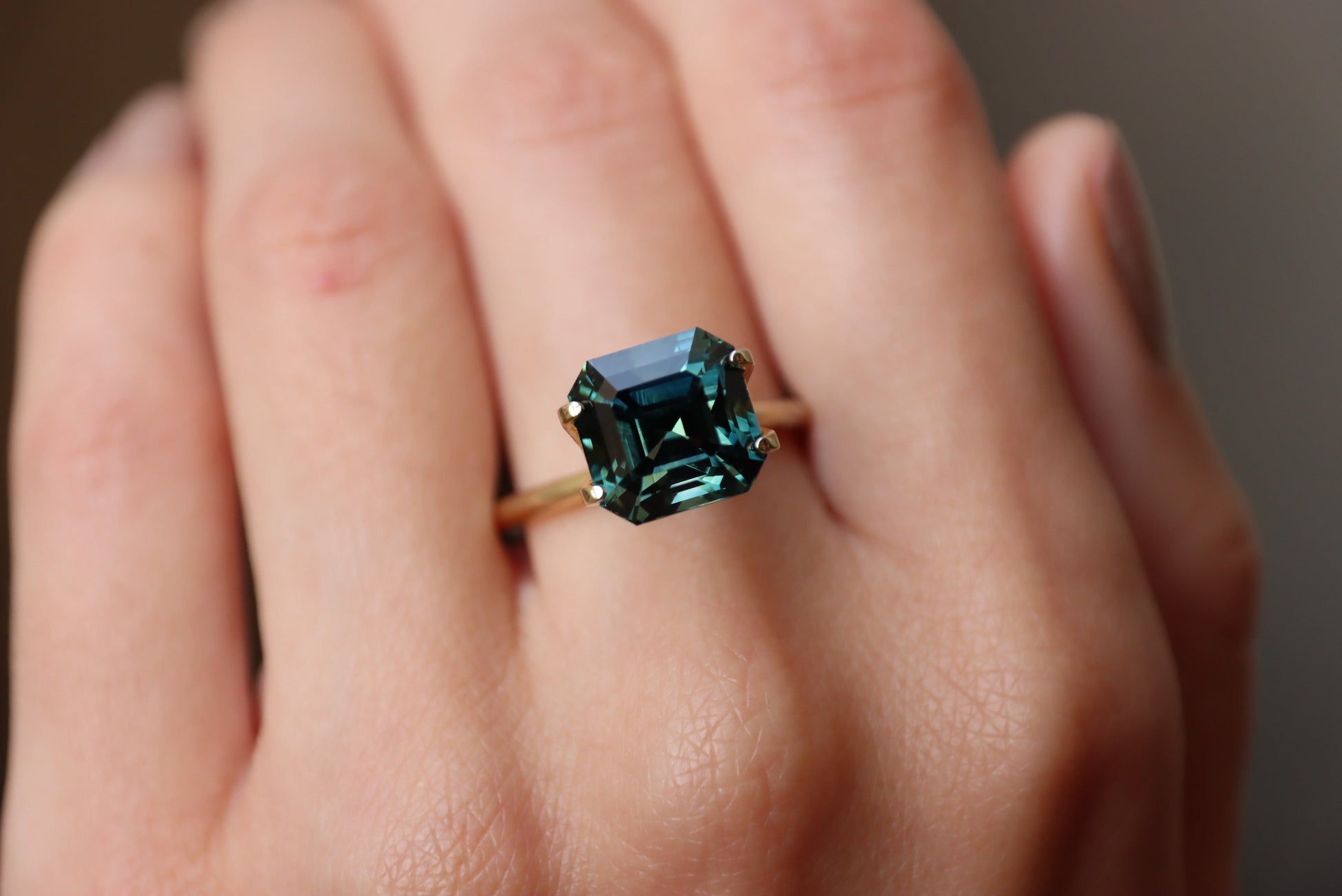 Une main met en valeur la bague en or Saphir Vert · 6,53 carats de Bonnot Paris, ornée d'une pierre turquoise taille émeraude sur un anneau lisse. Le flou de l'arrière-plan met en valeur les facettes détaillées de la bague.