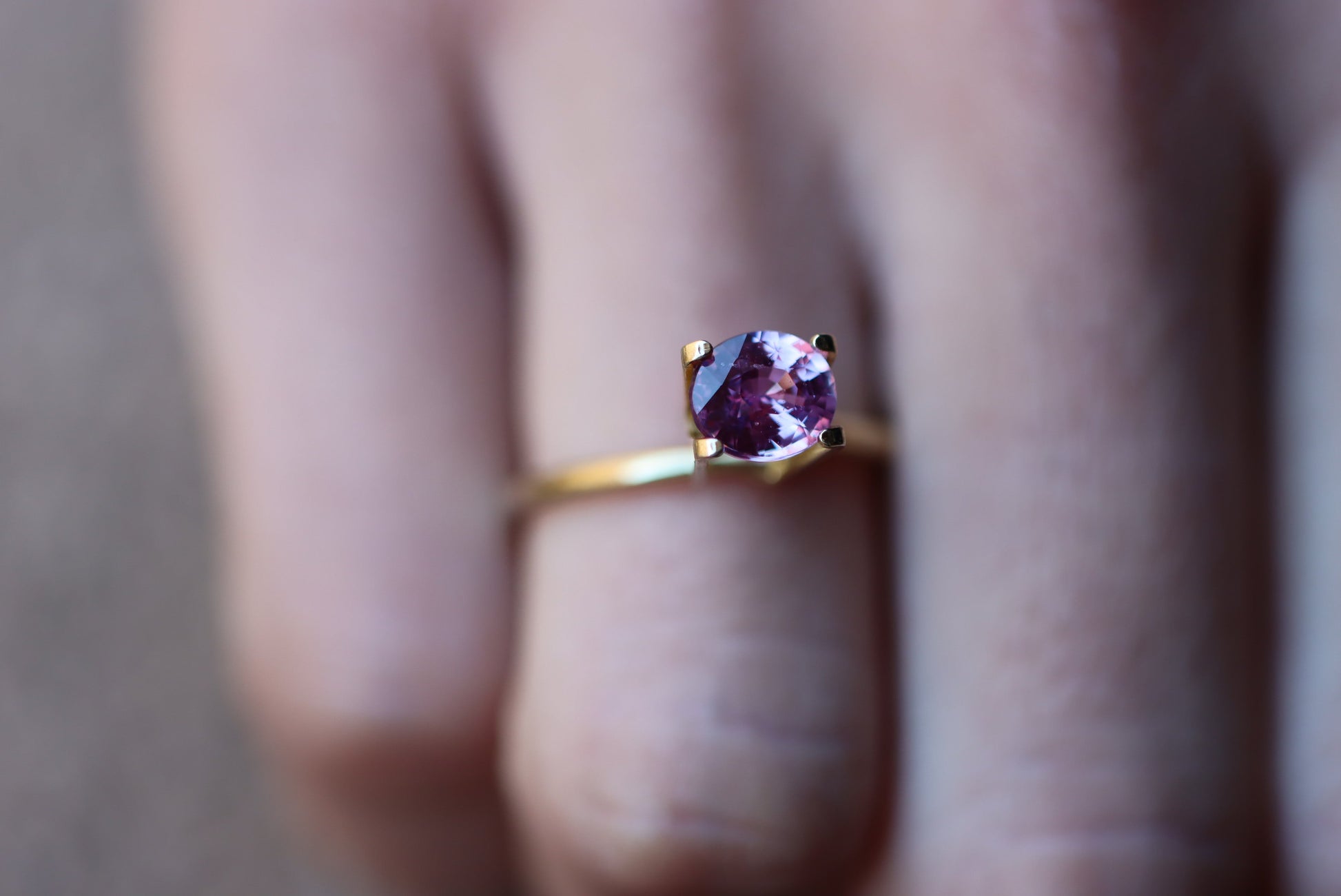 Un gros plan met en valeur une main portant la bague Saphir Violet en or 1,61 carat de Bonnot Paris, ornée d'une pierre violette à la taille scintillante, maintenue par quatre griffes. Elle se détache sur l'arrière-plan flou d'un doigt orné d'ongles neutres.