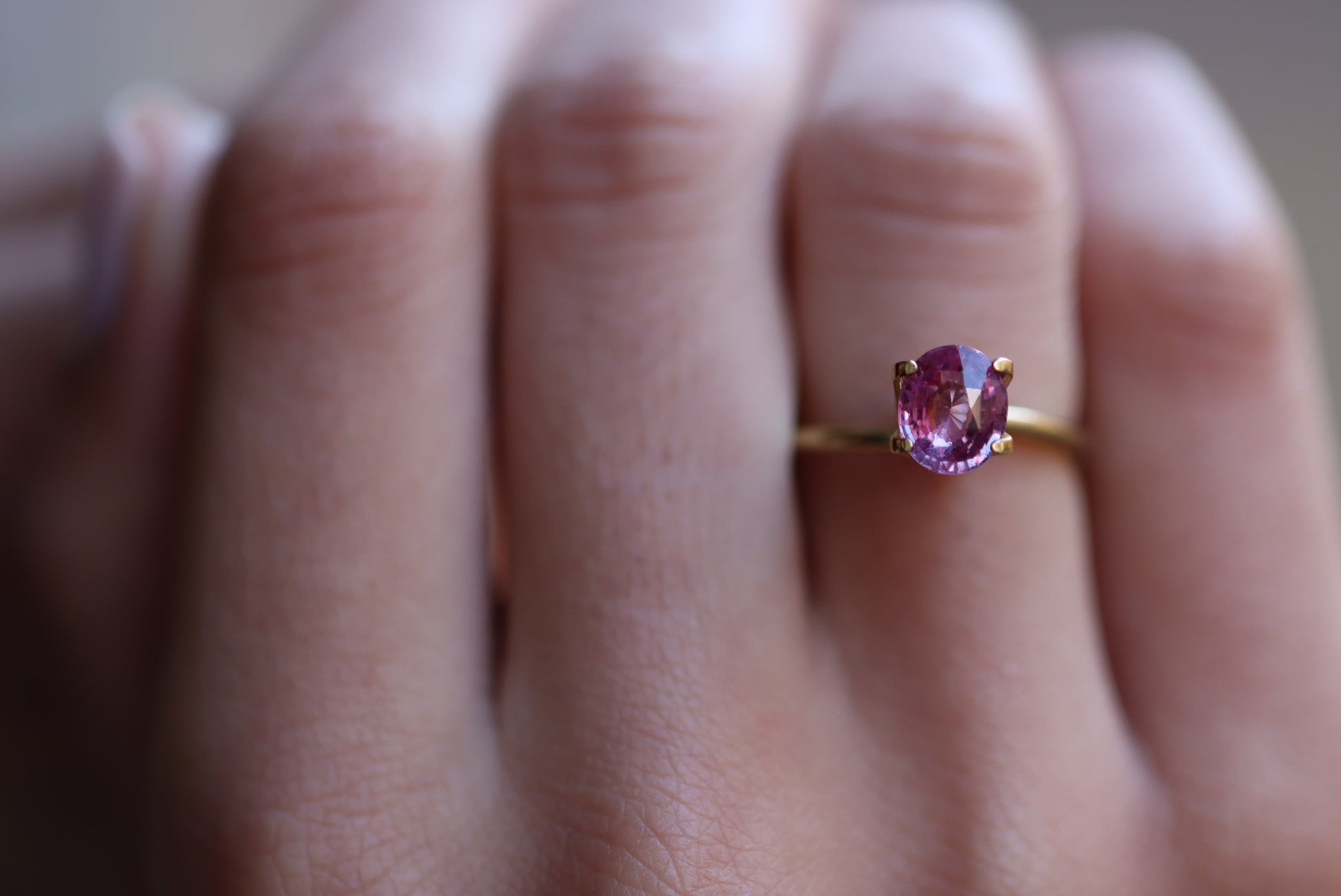 Gros plan sur une main ornée de la bague Saphir Violet 1,58 carat de Bonnot Paris, ornée d'une pierre rose ovale. La bague orne l'annulaire sur un fond légèrement flou, soulignant son élégance.