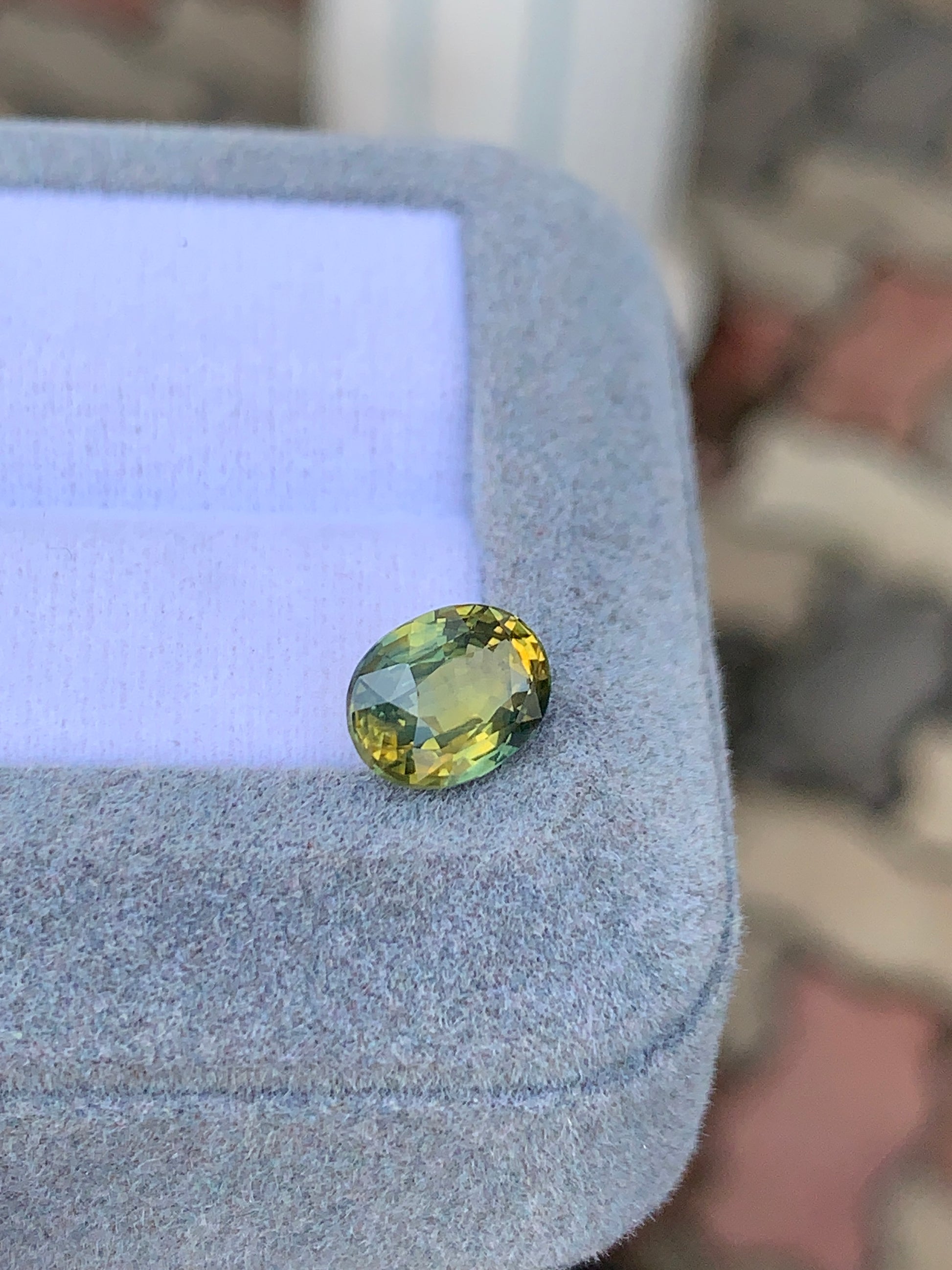 Une pierre précieuse Saphir Antique ovale étincelante, de 2,56 ct dans des teintes vertes et jaunes vibrantes, est élégamment placée sur une boîte à bijoux en velours gris clair de la collection [NOUVEAU] Bonnot Paris, sur un fond flou.