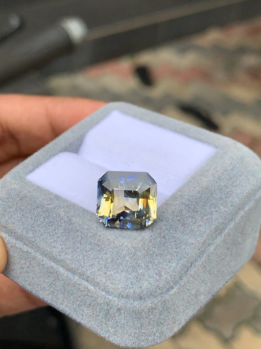 Une main tient une boîte à bague en velours gris ouverte contenant un Saphir Bicolore 10 carats de [NOUVEAU] Bonnot Paris, affichant une pierre précieuse rectangulaire avec un dégradé bleu et jaune fascinant sur des carreaux gris et marron flous.