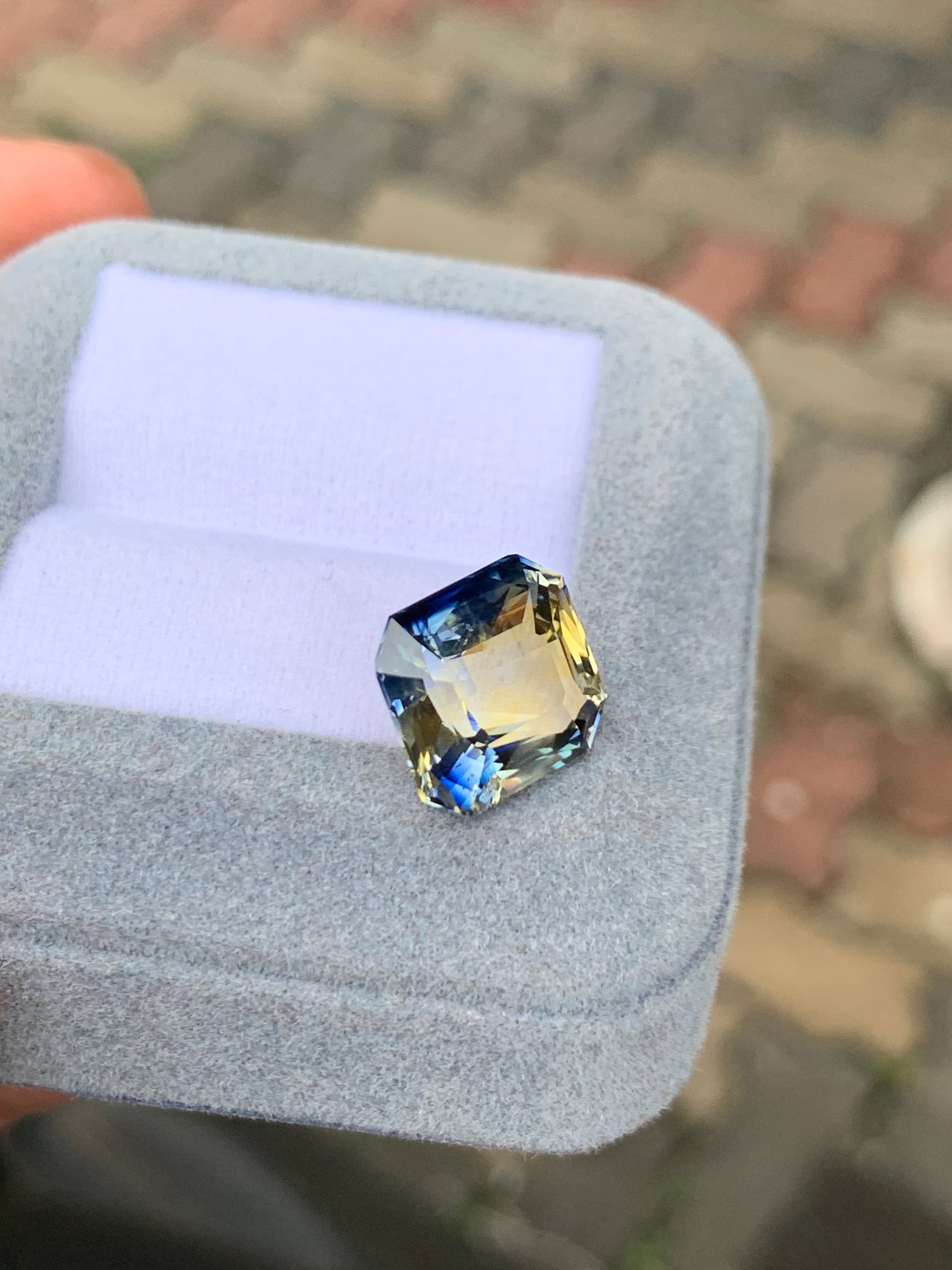 Une boîte à bijoux grise ouverte avec un intérieur violet clair abrite le [NOUVEAU] Bonnot Paris Saphir Bicolore • 10ct, une pierre précieuse aux teintes bleues et jaunes, tandis que l'arrière-plan affiche une surface pavée légèrement floue.
