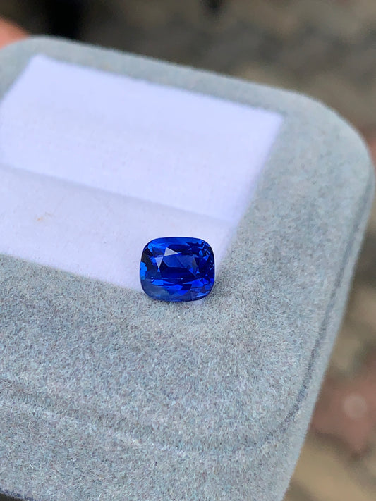 Un Saphir Bleu taille coussin bleu vif de 2,02 ct de Bonnot Paris est présenté dans une boîte en feutre gris clair avec un insert blanc, mettant en valeur la couleur profonde et les qualités réfléchissantes de la pierre précieuse.