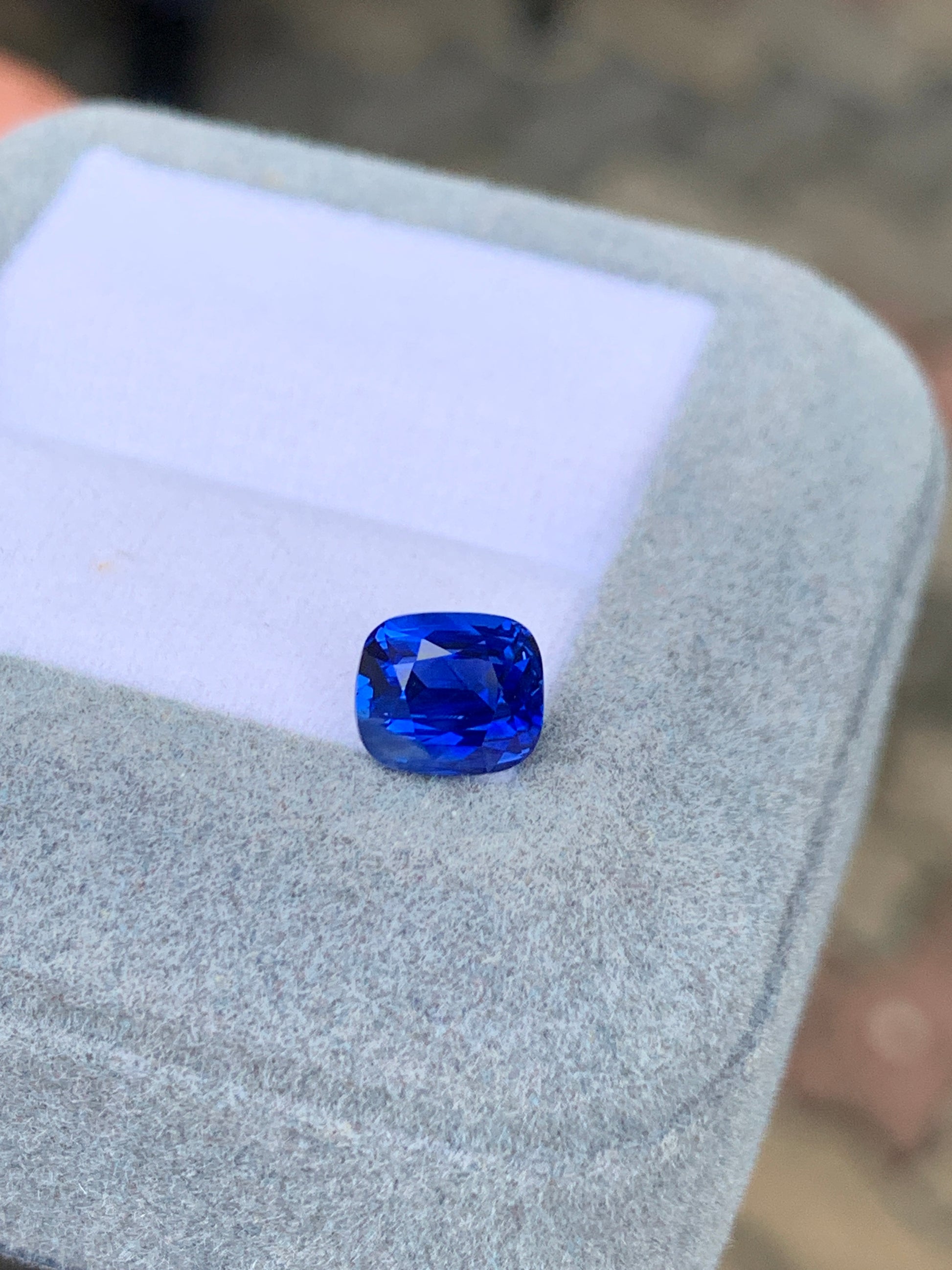 Le Saphir Bleu • 2,02ct de [NOUVEAU] Bonnot Paris, une pierre précieuse taille coussin d'origine sri-lankaise, est présenté dans une boîte gris clair sur un fond flou, mettant en valeur sa couleur vive et ses facettes, idéale pour une bague de fiançailles exquise.