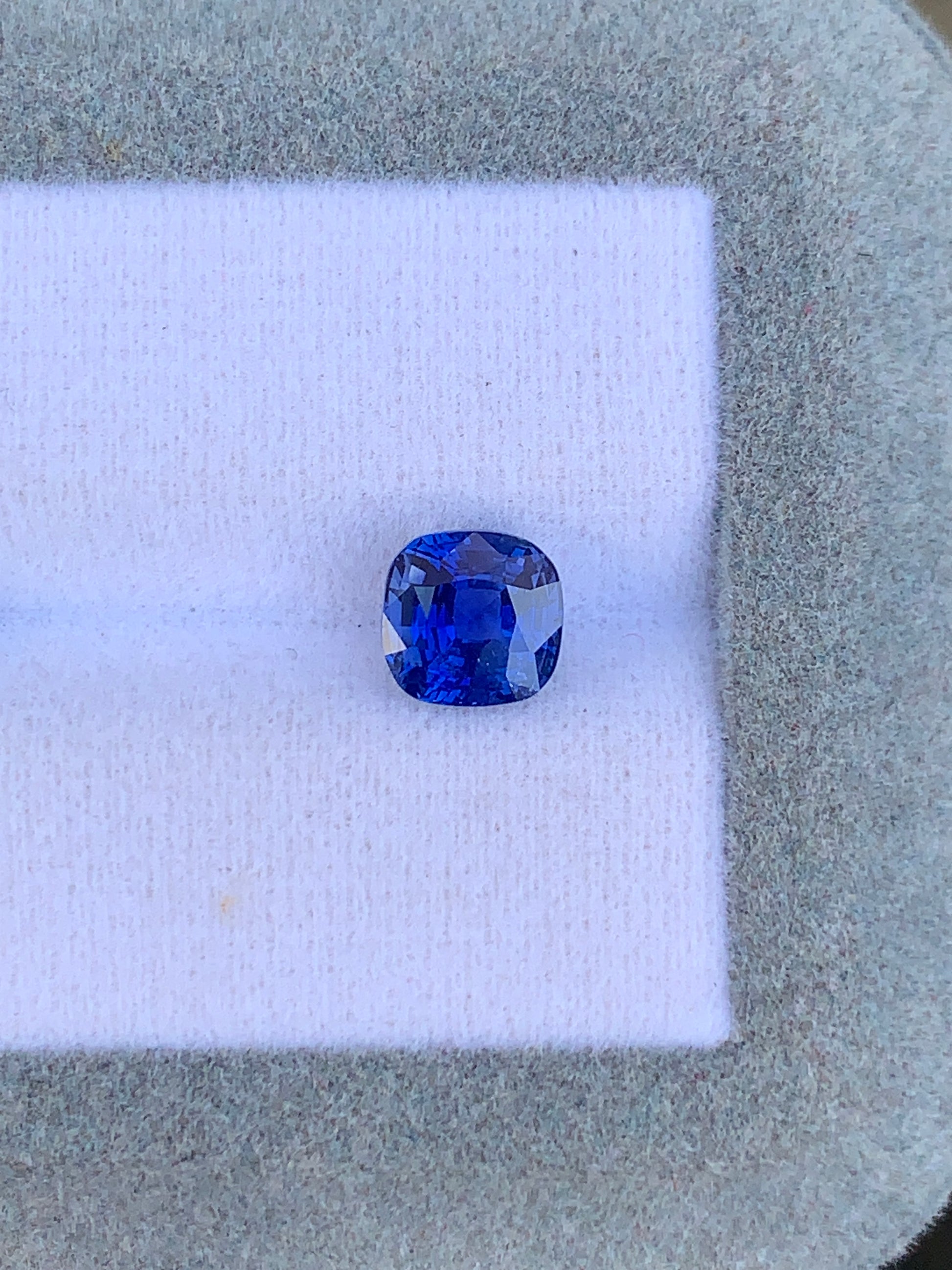 Une pierre précieuse Saphir Bleu taille coussin de 2,10 ct de Bonnot Paris scintille sur un fond de tissu blanc dans un cadre texturé gris, affichant sa teinte bleue vibrante - une véritable création joaillière.