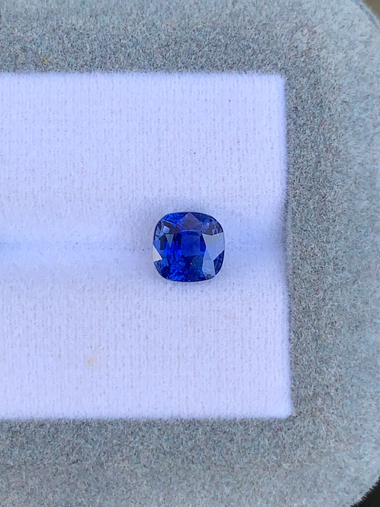 Une pierre précieuse Saphir Bleu taille coussin de 2,10 ct de Bonnot Paris scintille sur un fond de tissu blanc dans un cadre texturé gris, affichant sa teinte bleue vibrante - une véritable création joaillière.