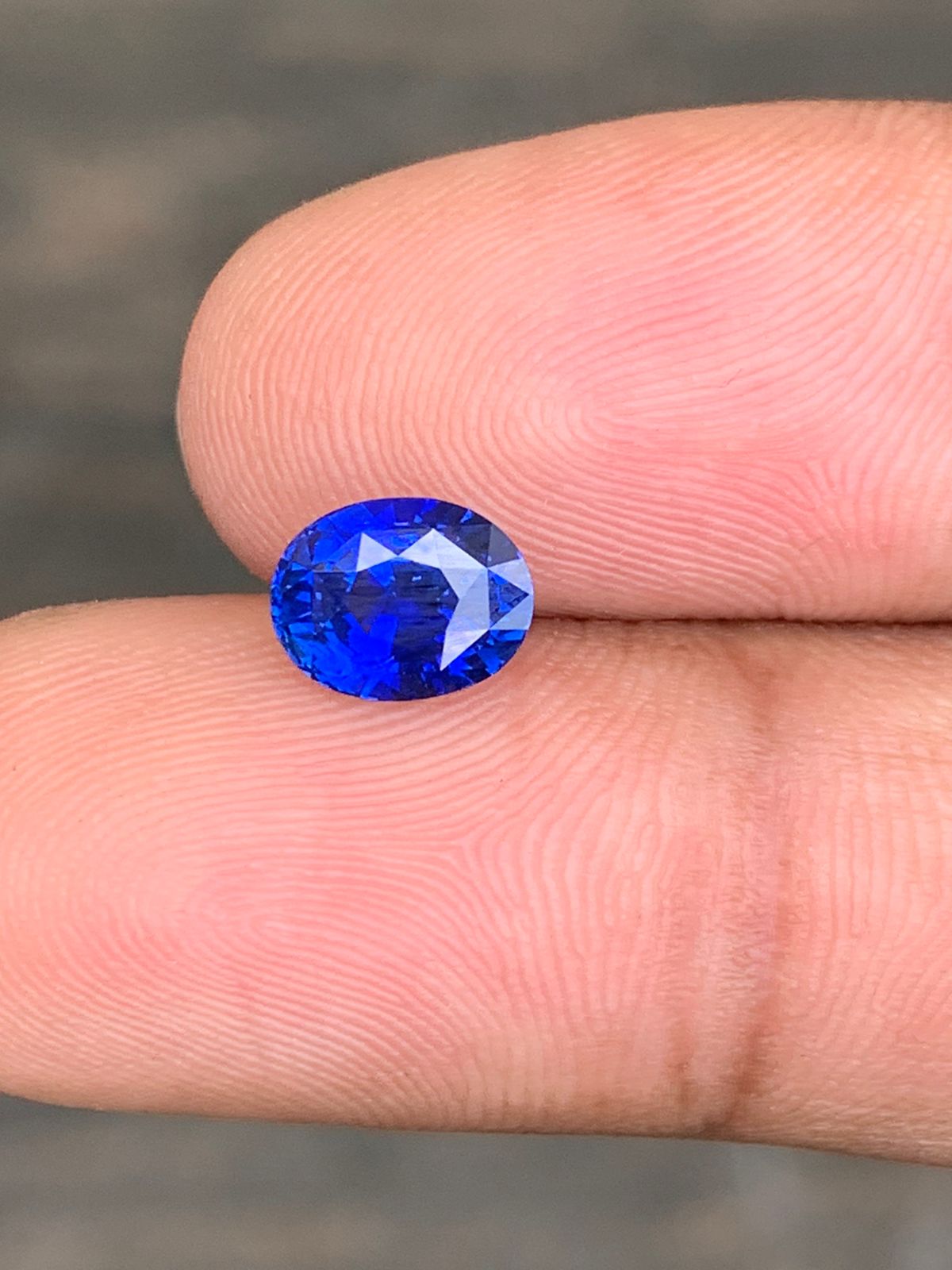 Le Saphir Bleu • 2,2ct de Bonnot Paris est délicatement tenu entre le pouce et l'index sur un fond flou, ses facettes bleues vives reflétant la lumière pour mettre en valeur sa clarté et sa couleur vibrante.