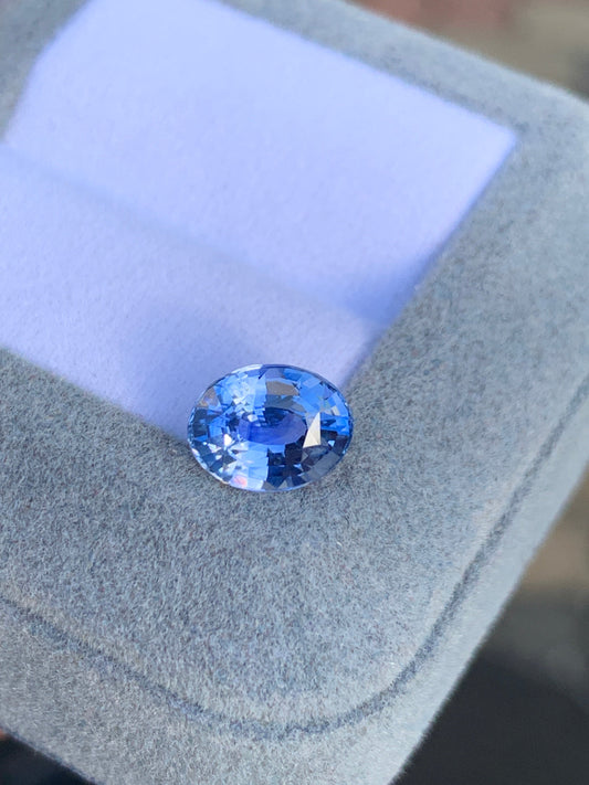 Le Saphir Bleu • 2,60ct de Bonnot Paris, une pierre précieuse bleue brillante à facettes, repose sur un coffret gris doux doublé de tissu violet clair, reflétant la lumière et mettant en valeur sa couleur vibrante et sa clarté.