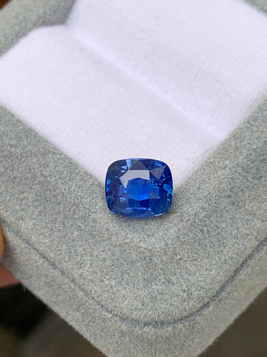 Le Saphir Bleu Bonnot Paris de 3,04 ct, en provenance du Sri Lanka, met en valeur ses éclatantes nuances bleu bleuet et sa taille coussin sur une surface grise texturée, à l'intérieur d'un écrin partiellement visible doublé de tissu blanc. Cette pierre précieuse non chauffée scintille à la lumière, révélant sa couleur riche et ses magnifiques facettes.
