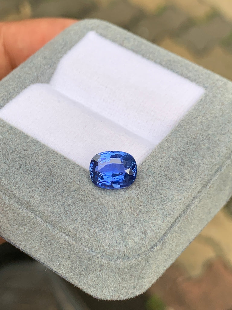 Un gros plan du Saphir Bleu · 3,05ct de [NOUVEAU] Bonnot Paris sur une boîte à bijoux gris clair avec un intérieur blanc met en valeur la couleur vibrante de la pierre précieuse taille coussin sur un fond flou.