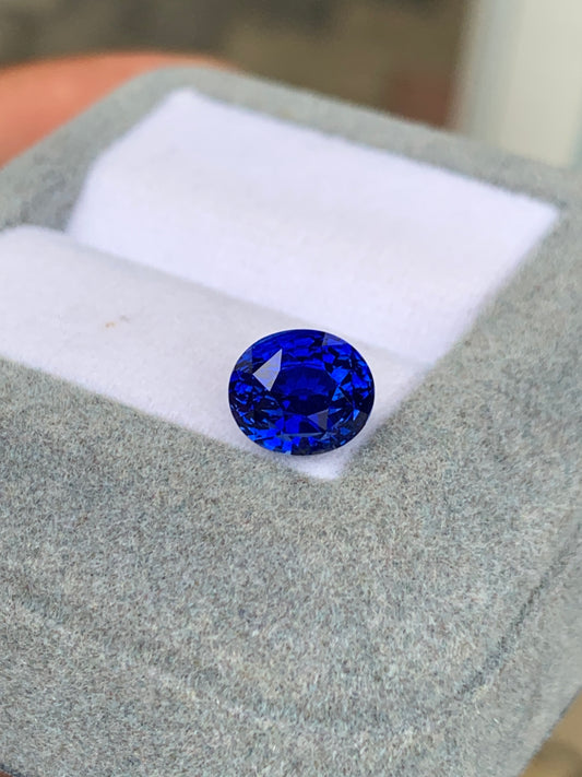 Le Saphir Bleu • 3,25ct de Bonnot Paris est une pierre précieuse ovale, d'un bleu vibrant, aux multiples facettes, présentée dans un écrin à bijoux blanc matelassé sur un extérieur en tissu gris.