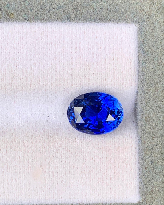 Un Saphir Bleu vibrant • 4,15 ct de [NOUVEAU] Bonnot Paris, taille ovale et provenant du Sri Lanka, est élégamment présenté sur un tissu rose clair doux sur un fond gris pâle texturé.