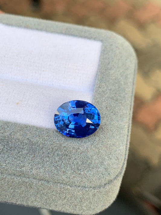 Le Saphir Bleu • 5 carats de Bonnot Paris, une pierre précieuse de taille ovale intense, repose sur une surface coussinée gris clair. Sa teinte saphir profonde rappelle celle des saphirs du Sri Lanka et est inclinée pour accentuer ses facettes et sa clarté sur un fond flou aux tons chauds.