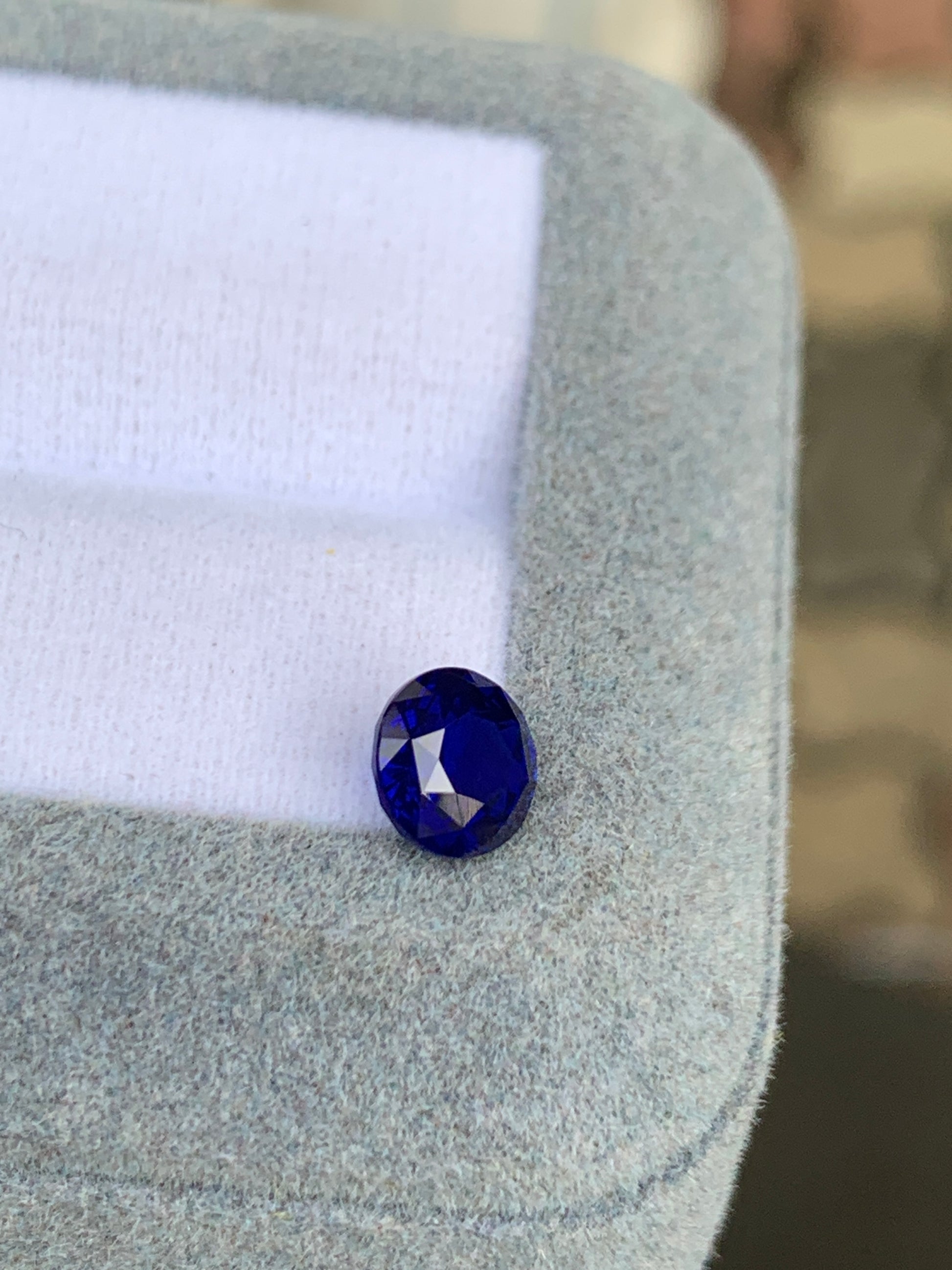 Un Saphir Bleu de 1,52 ct de [NOUVEAU] Bonnot Paris, une exquise pierre précieuse ovale du Sri Lanka, repose sur un tissu doux gris et blanc, avec sa couleur vive et ses facettes mises en valeur sur un fond flou.