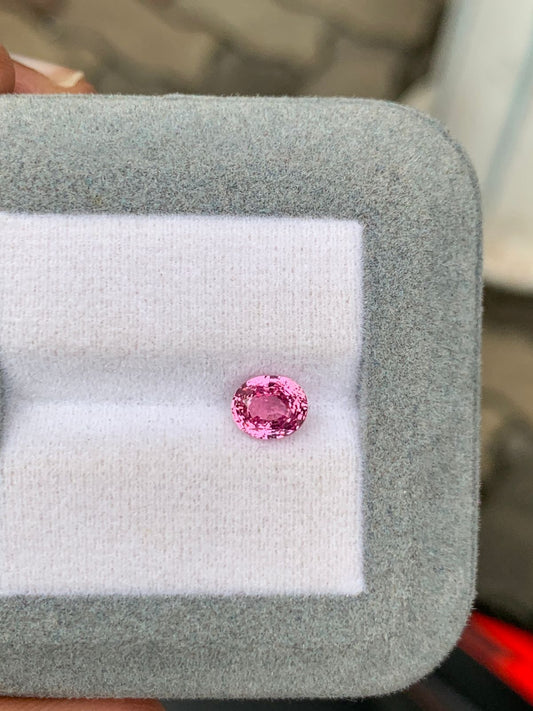 Une pierre précieuse ovale rose de 1,50 carat, baptisée Saphir Padparadscha par Bonnot Paris, repose élégamment sur un coussin blanc. Cette magnifique pierre est sertie dans un élégant cadre rectangulaire gris, saisissant la beauté naturelle de Madagascar.