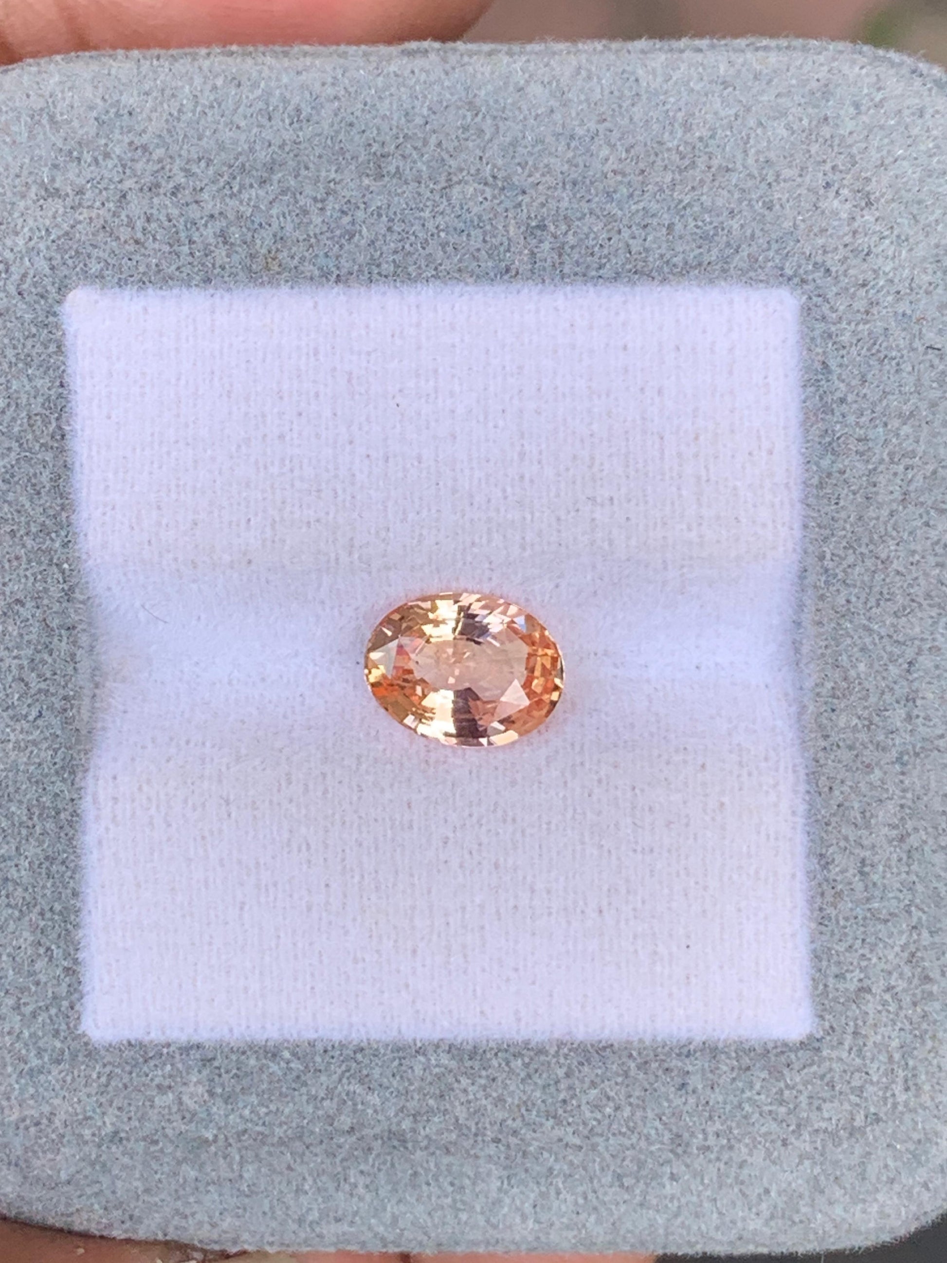 Un Saphir Padparadscha rond et facetté (2,04 carats) de Bonnot Paris est présenté sur un tissu blanc dans un support bleu-gris clair. Le fond flou met en valeur cette pierre précieuse exquise.