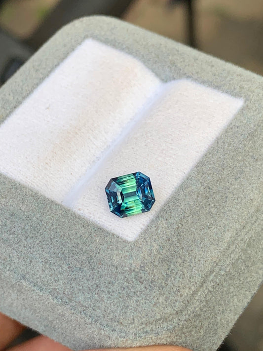 Le Saphir Teal de Bonnot Paris est une pierre précieuse taille émeraude scintillante de 1,65 carat. Logée dans un écrin doublé de velours gris, ses facettes révèlent des teintes captivantes allant du bleu profond au vert.