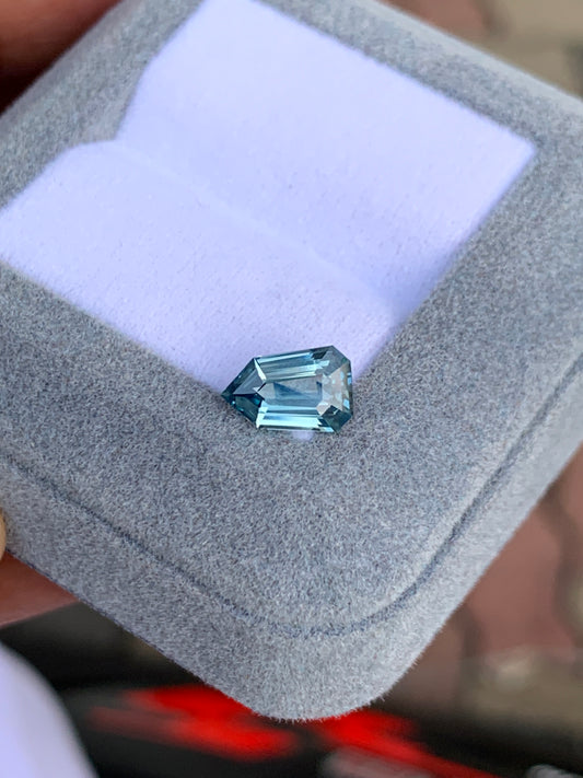 Le Saphir Teal de 1,65 carat de Bonnot Paris est présenté dans un écrin gris clair à l'intérieur blanc. Sa forme asymétrique aux multiples surfaces réfléchissantes met en valeur sa clarté et sa couleur sur un fond flou.