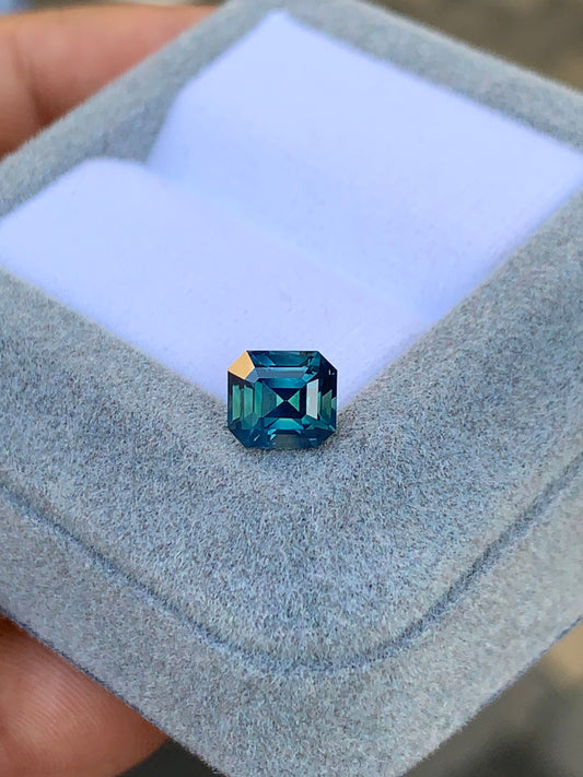 Une main tient une boîte à bijoux grise avec un intérieur coussiné, mettant en vedette le Saphir Teal • 1,70 ct de Bonnot Paris, une pierre précieuse bleu-vert foncé taille émeraude.
