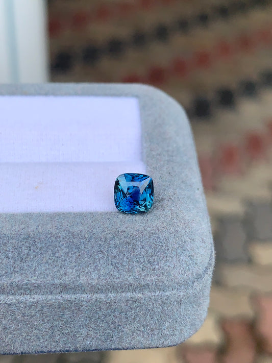Le Saphir Teal • 2,05 carats de Bonnot Paris, une pierre précieuse éclatante, présente une taille coussin sur une surface grise texturée. Le fond légèrement flou met en valeur la richesse de la couleur et des facettes de cette pierre rare.