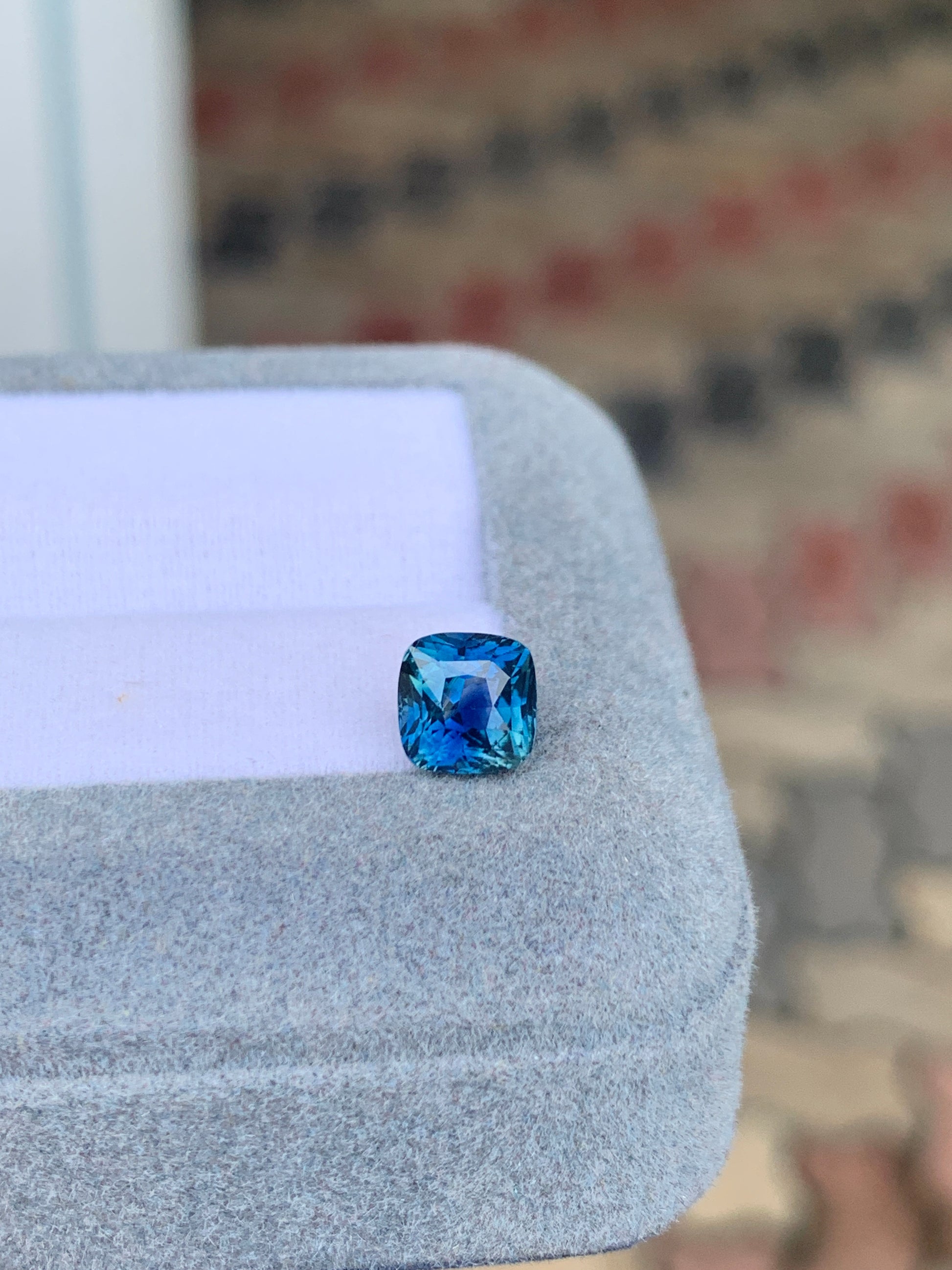 Le Saphir Teal • 2,05 ct de [NOUVEAU] Bonnot Paris, une pierre taille coussin, est posé sur un écrin gris sur un fond flou aux motifs de briques. Il éblouit par ses nuances bleues et bleu sarcelle, révélant son éclat facetté.