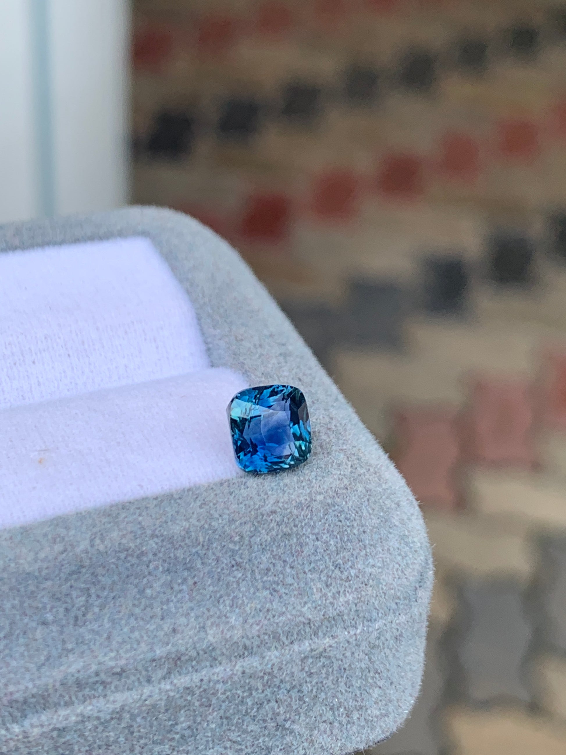 Le « Saphir Teal • 2,05 ct » de [NOUVEAU] Bonnot Paris, une pierre précieuse bleu-vert taille carrée, repose sur un coffret en velours gris sur un fond de pavés légèrement flous.