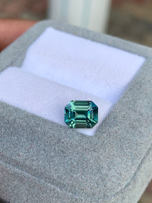 Les doigts d’une personne arborent le Saphir Teal • 2,01ct de [NOUVEAU] Bonnot Paris : une pierre précieuse verte taille émeraude, non chauffée et réfléchissant la lumière de ses facettes précises, sertie sur un écrin à bijoux gris avec une doublure blanche.