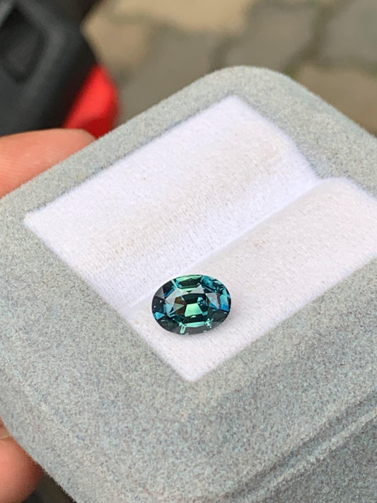 La pierre précieuse Saphir Teal 2,03 ct, de [NOUVEAU] Bonnot Paris, présente des teintes bleu et vert profonds, élégamment serties dans un coffret à bijoux gris avec une doublure blanche immaculée.