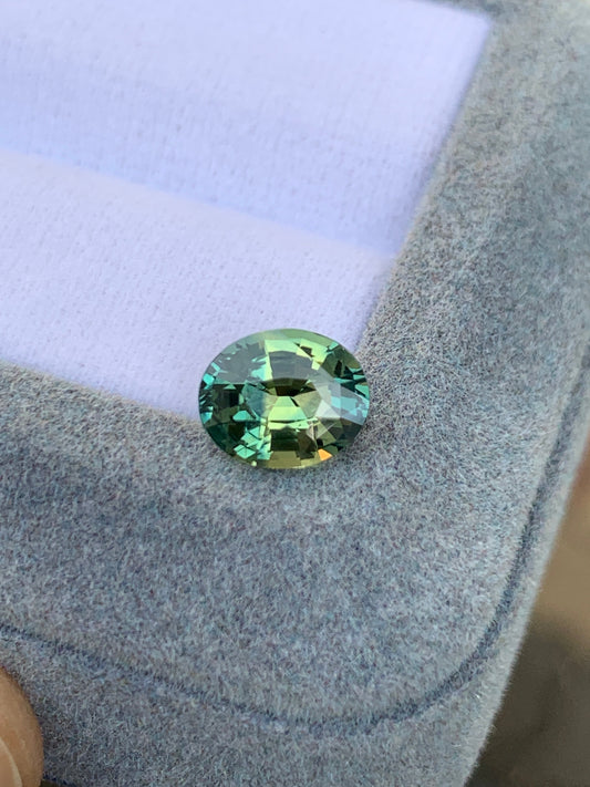 Une pierre précieuse ronde Saphir Teal pesant 2,04 ct, présentant un mélange multicolore de teintes vertes et jaunes, est présentée sur un tissu gris clair de Bonnot Paris.