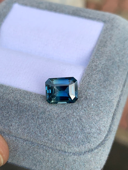 Le Saphir Teal • 2,40ct de Bonnot Paris, une pierre précieuse taille émeraude d'un bleu profond, repose sur un écrin à bijoux gris clair avec un intérieur blanc.
