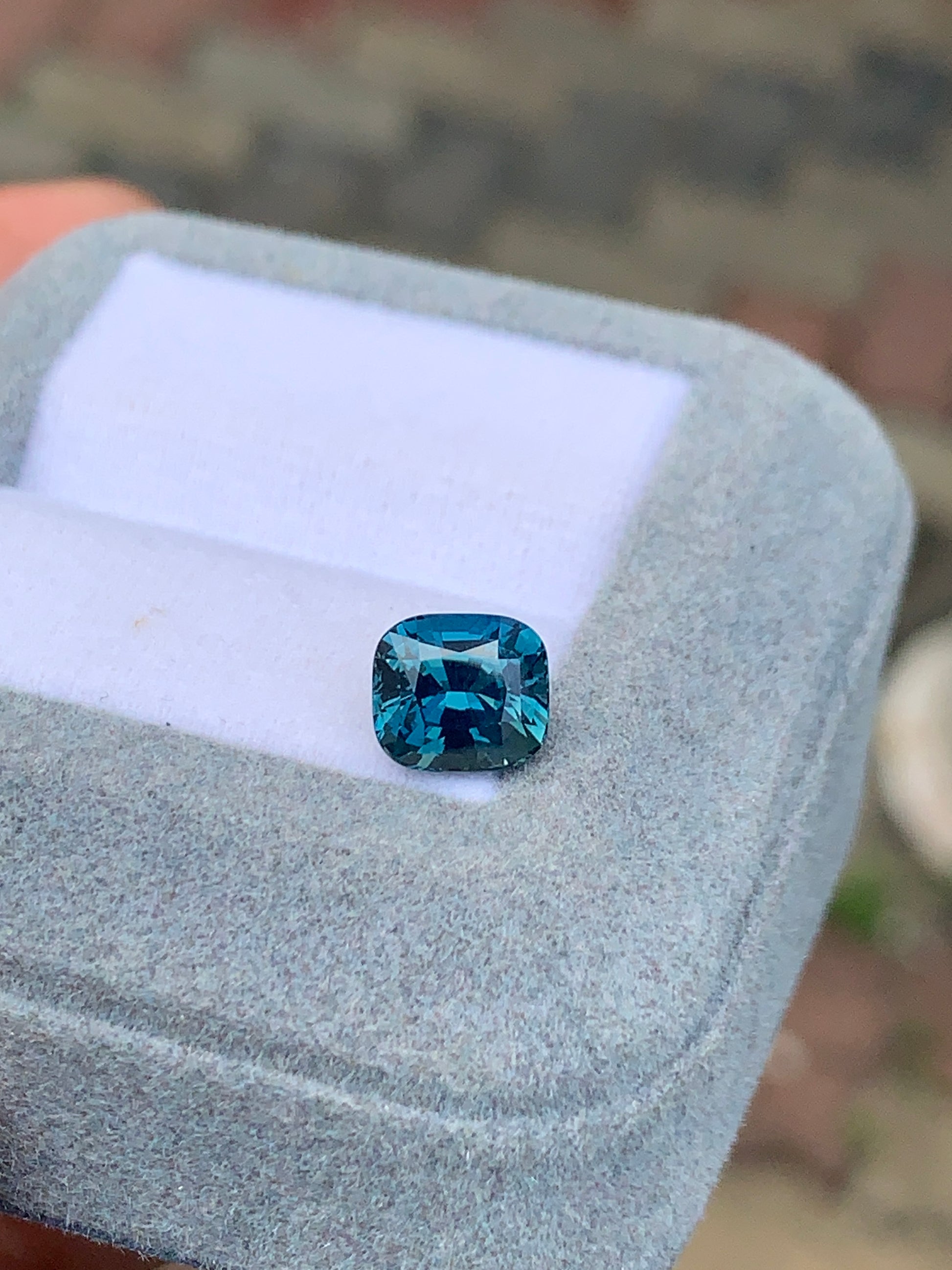 Le Saphir Teal • 2,50 carats de Bonnot Paris, un saphir taille coussin de 2,50 carats, est élégamment présenté dans un écrin de velours gris clair. Un fond légèrement flou met en valeur les détails exquis de ce trésor de Madagascar sur une surface aux motifs subtils.