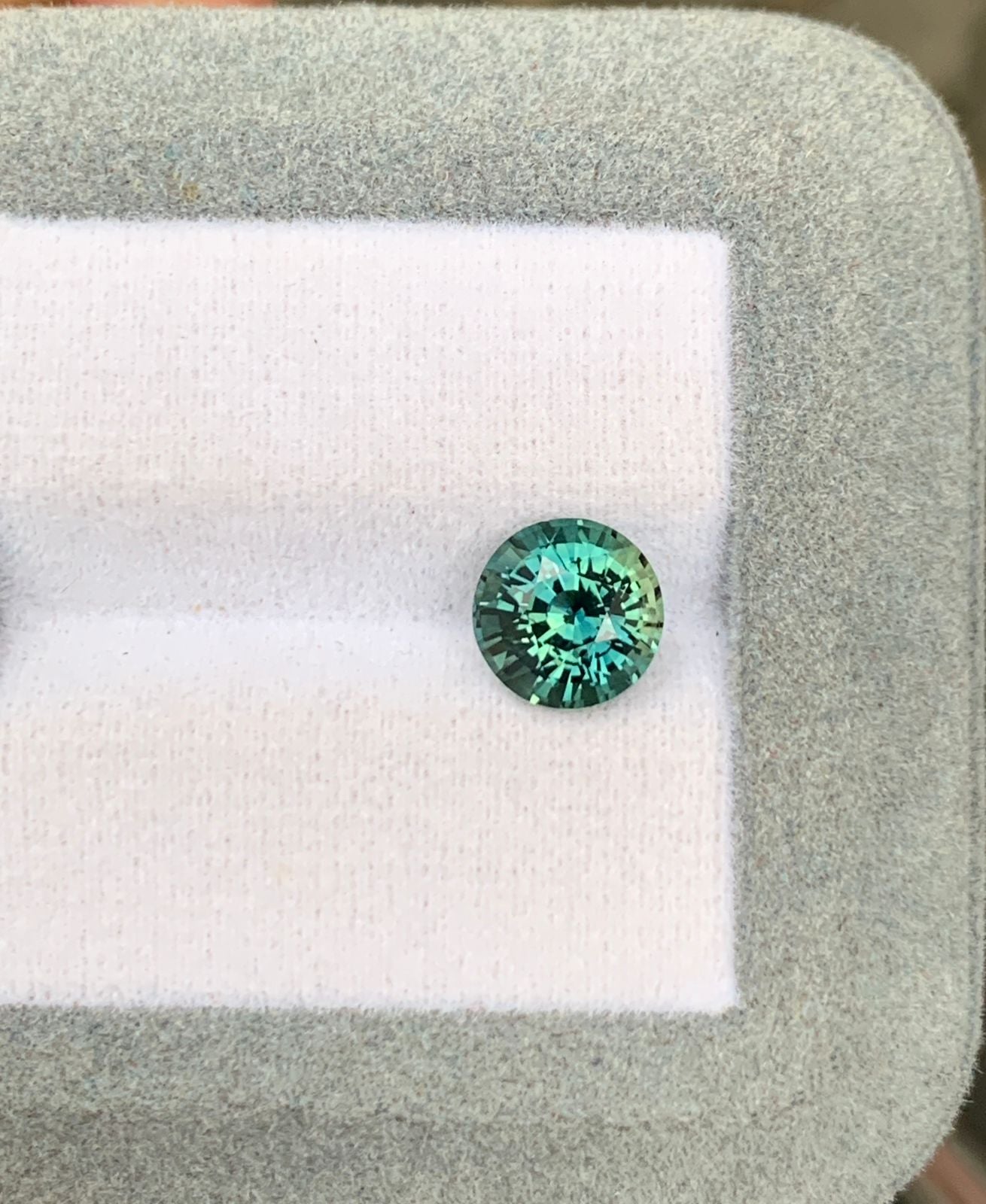Une pierre précieuse Saphir Teal étincelante de 2,52 ct de [NOUVEAU] Bonnot Paris de Madagascar repose sur une plate-forme coussinée blanche et grise, affichant ses facettes bleu-vert vibrantes avec un fond doucement flou.